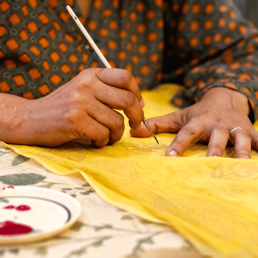 An artisan uses a fine brush to carefully paint intricate designs onto bright yellow cloth.