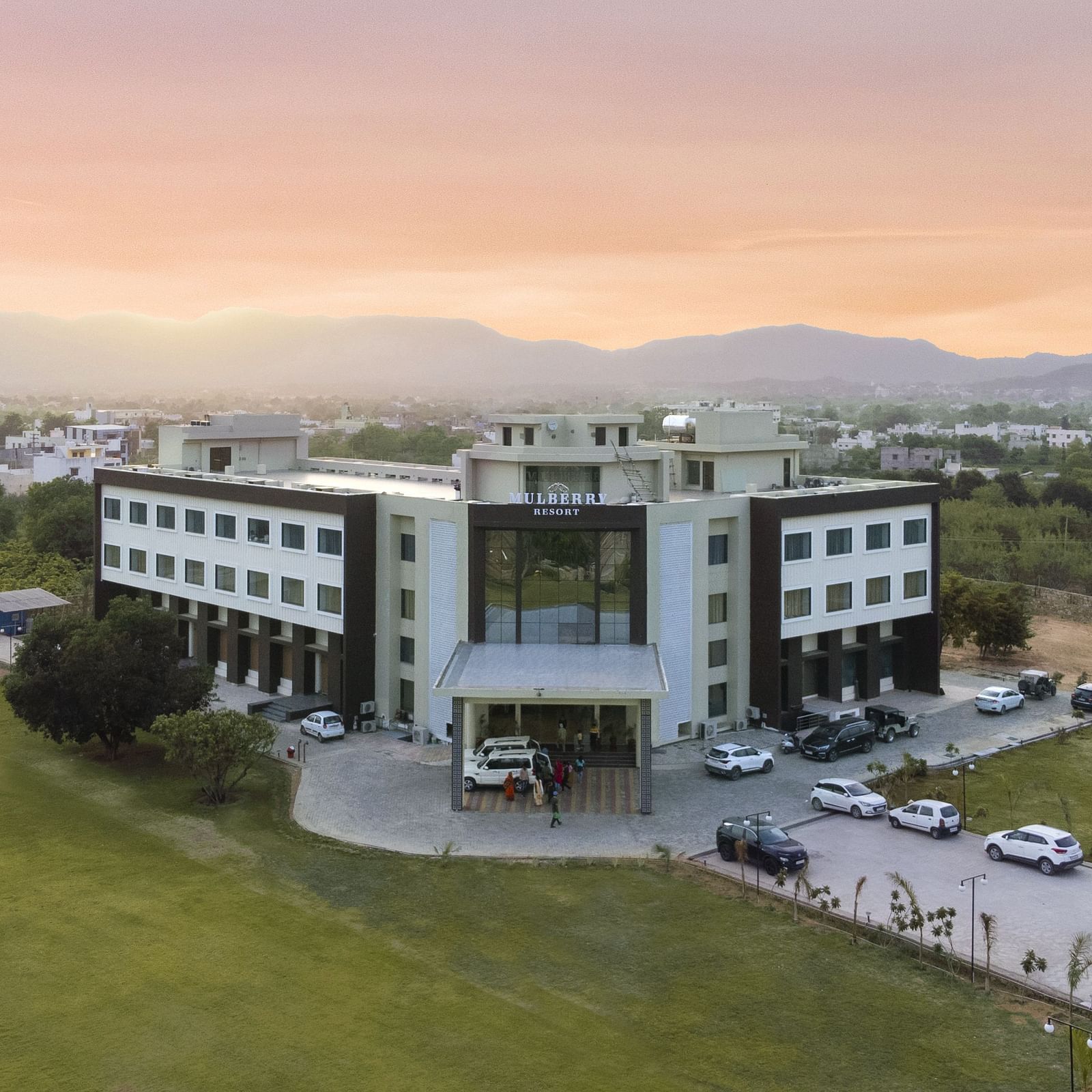 An aerial view of the Bamboo Saa Mulberry Resort, Pushkar at sunset or sunrise surrounded by green lawn and the backdrop of hills.