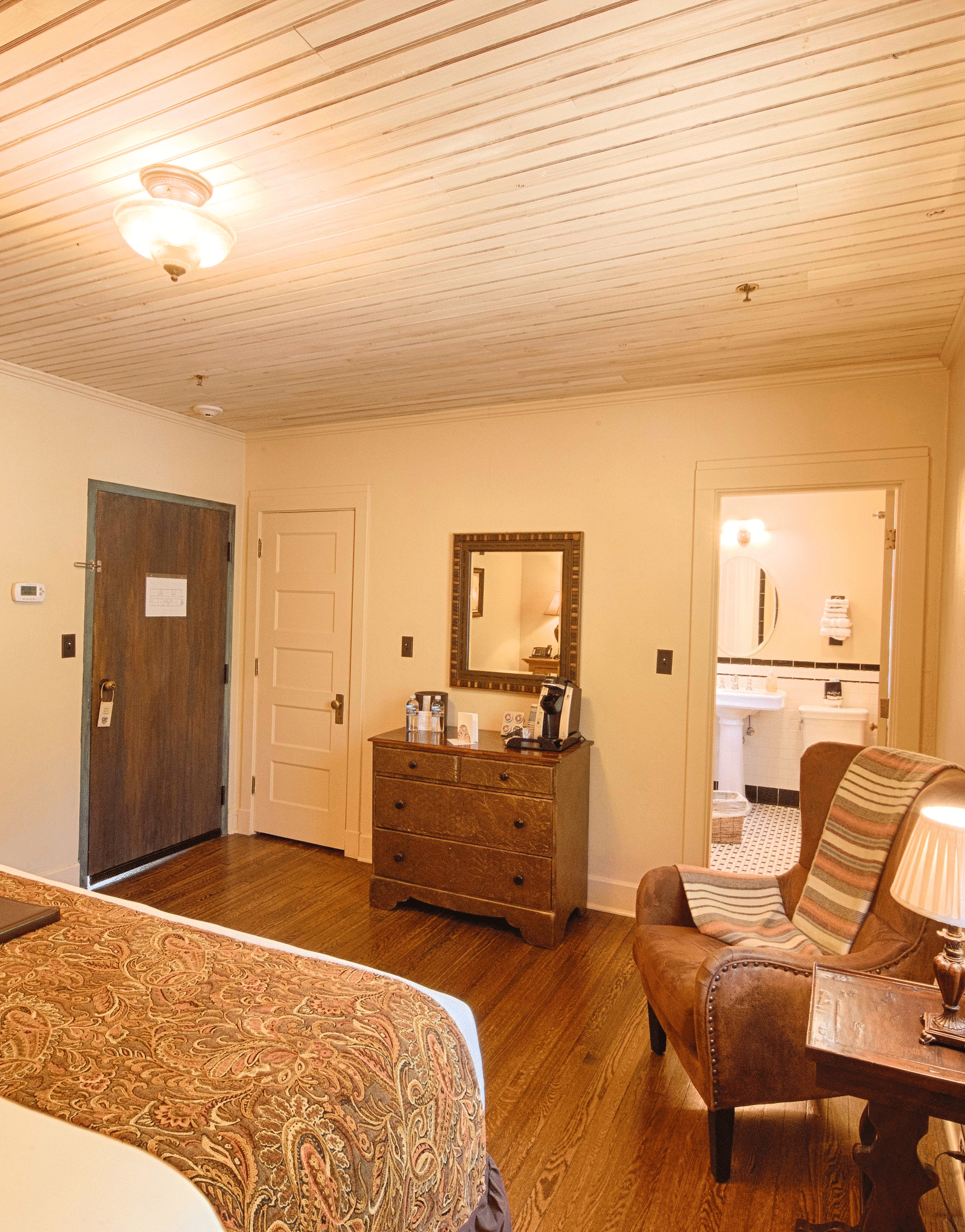 a cozy bedroom with a cushioned rustic armchair along with a small side table with a table lamp and a double bed in the room - Historic Tapoco Lodge