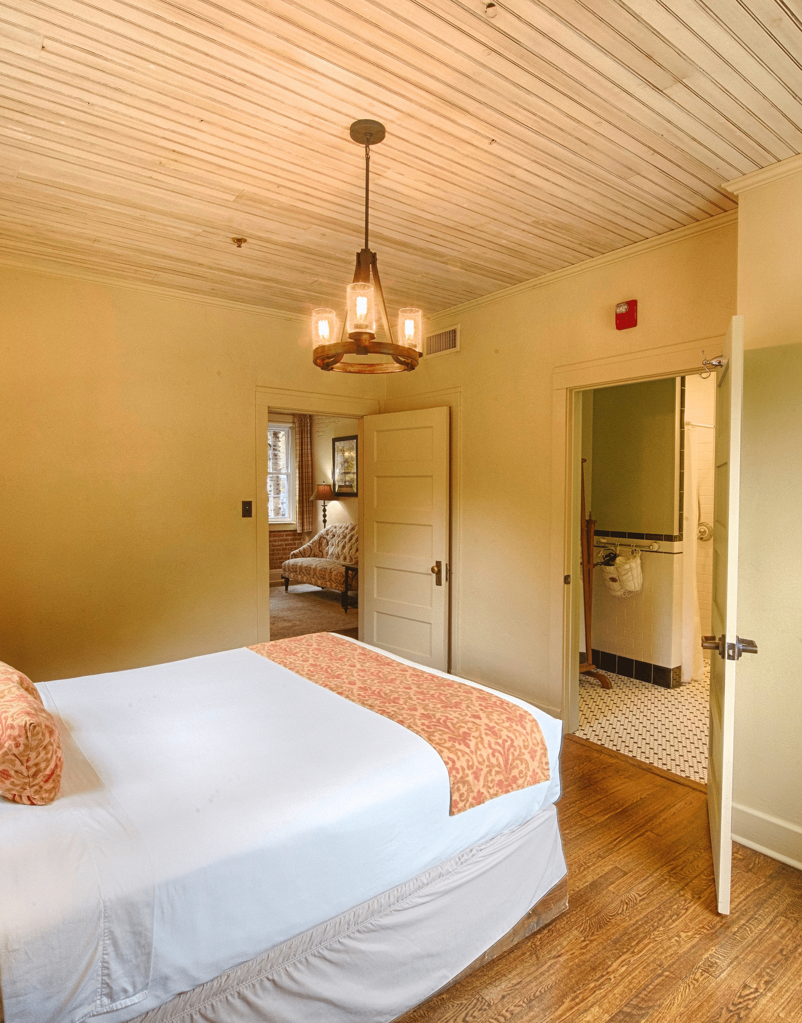 a room with two door along with a double bed and a chandelier hanging over ita brightly lit bathroom featuring a door opening to the bedroom overlooking the double bed - Historic Tapoco Lodge