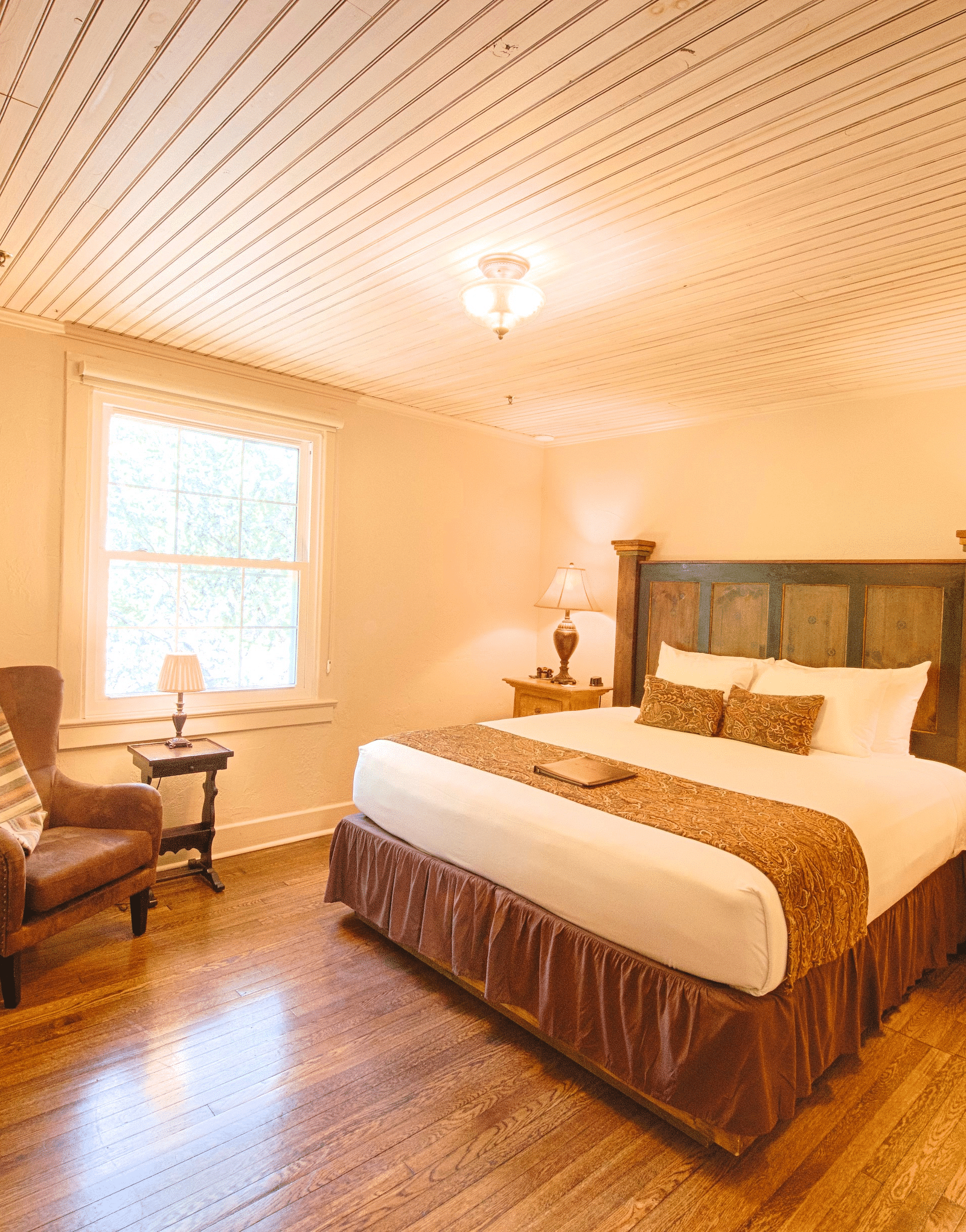 full view image of a brightly lit bedroom with a comfortable double bed with a heady headboard along with an armchair - Historic Tapoco Lodge