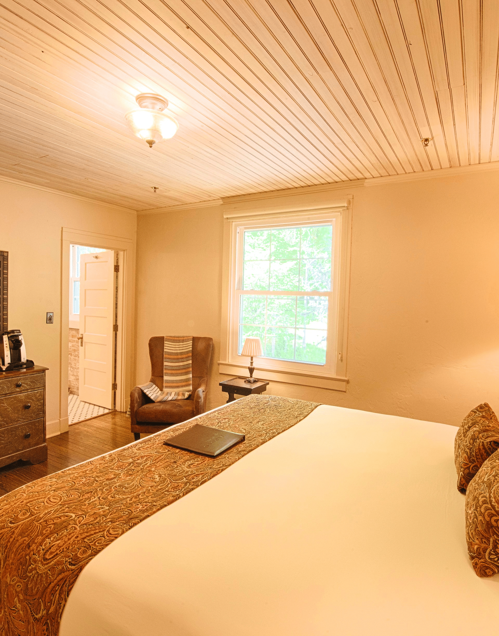 corner view image of a bedroom featuring a plush soft double bed with a small window in the far end of the room - Historic Tapoco Lodge