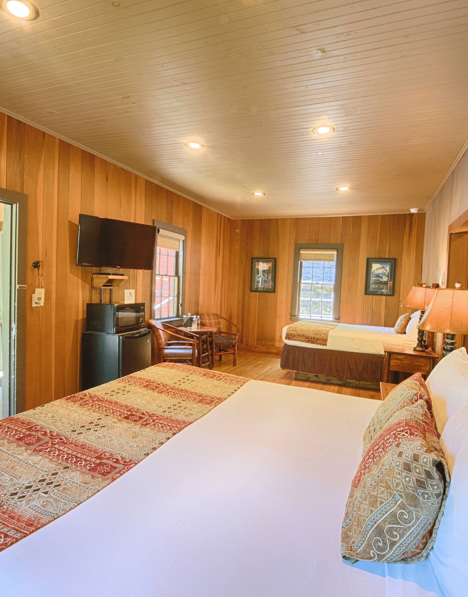 a room with wooden walls featuring two double beds with plush pillows and cushions along with a wall mounted television - Historic Tapoco Lodge