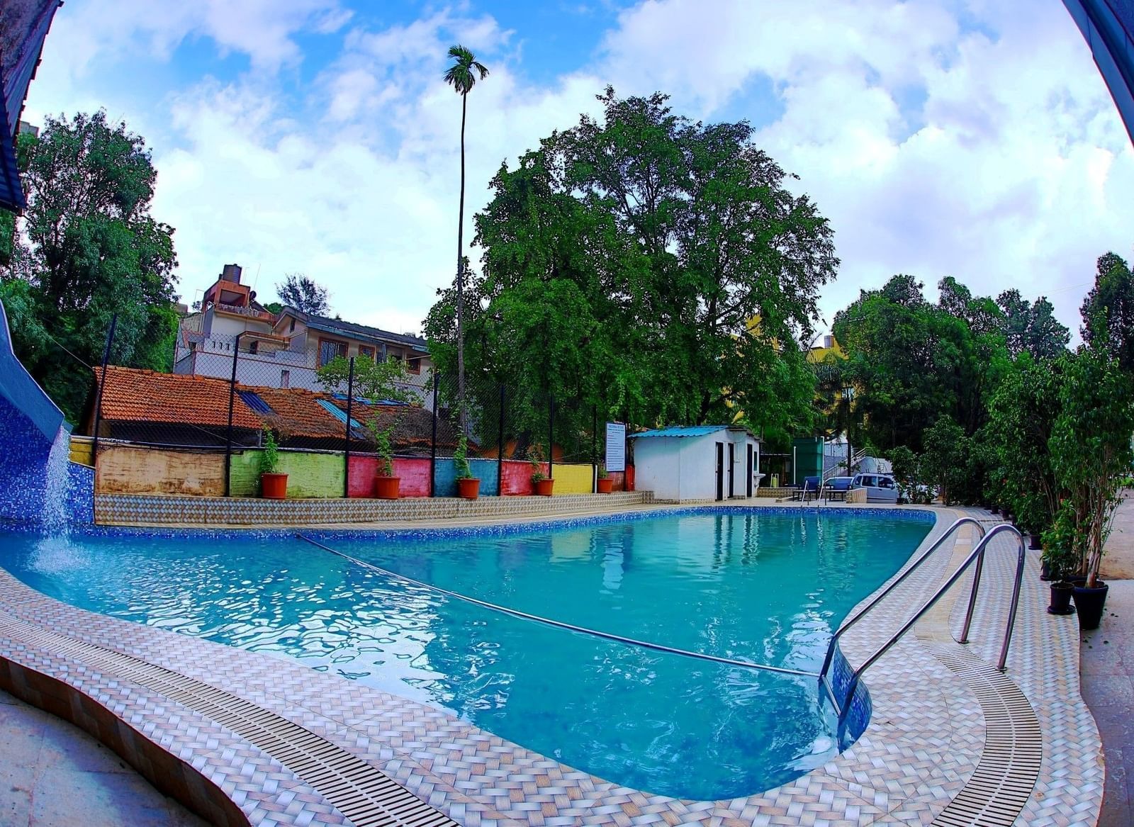 Assymetrical large swimming pool at Krushnai Resort under a partly cloudy blue sky.