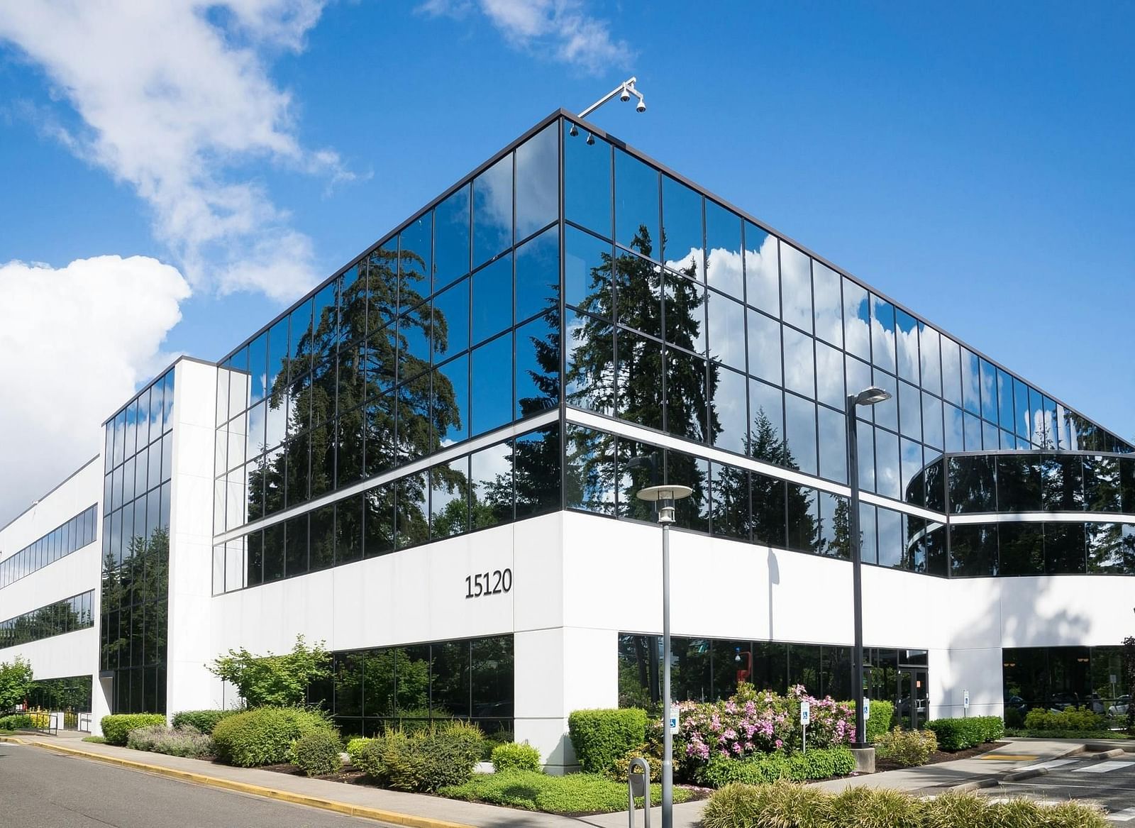 A modern, angular office building with glass walls reflecting the blue sky and surrounding greenery.