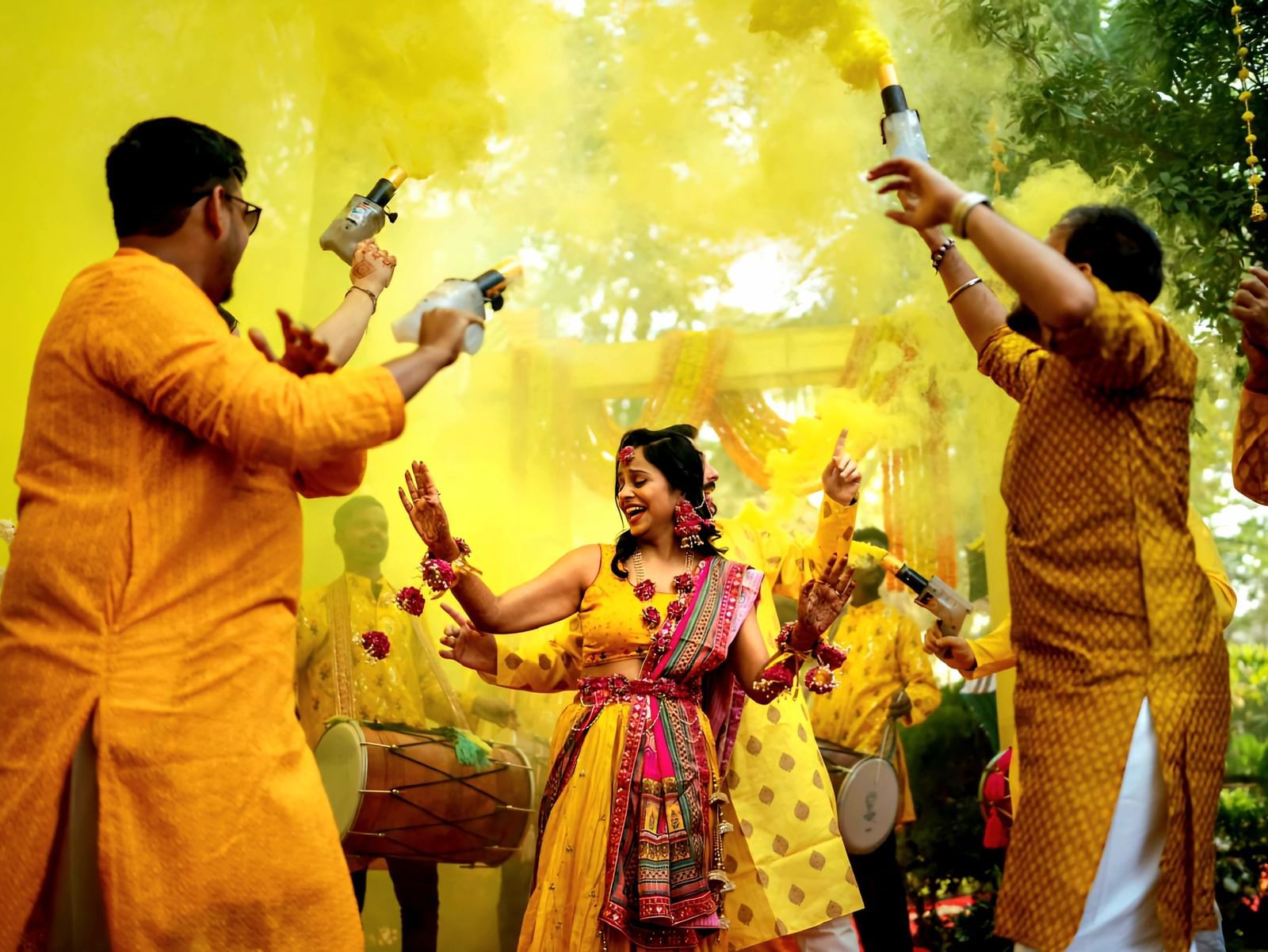 Two men in traditional attire pour a substance over a woman during a vibrant Indian ceremony, possibly a Haldi ceremony