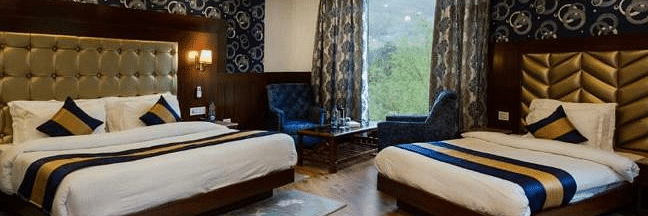 A spacious, well-lit hotel room at La Serene Valley by DLS Hotels in Manali, India. The room features two beds, a sitting area, and large windows offering natural light and views of the surrounding mountains | DLS La Serene Manali