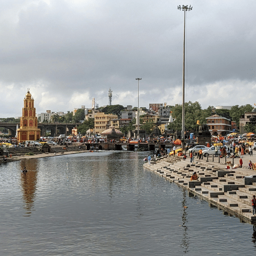 An aerial view of a bathing ghat in Nashik with the Godavari River in view, many people near the steps of the river and dark clouds above.