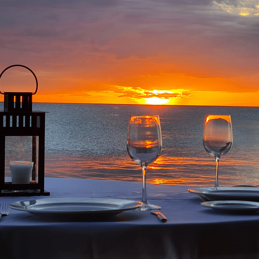 Romantic private dinner table for two with wine glasses and a lantern on the beach during a vibrant orange sunset