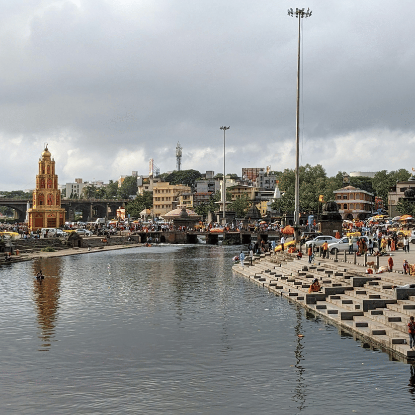 An aerial view of a bathing ghat in Nashik with the Godavari River in view, many people near the steps of the river and dark clouds above.