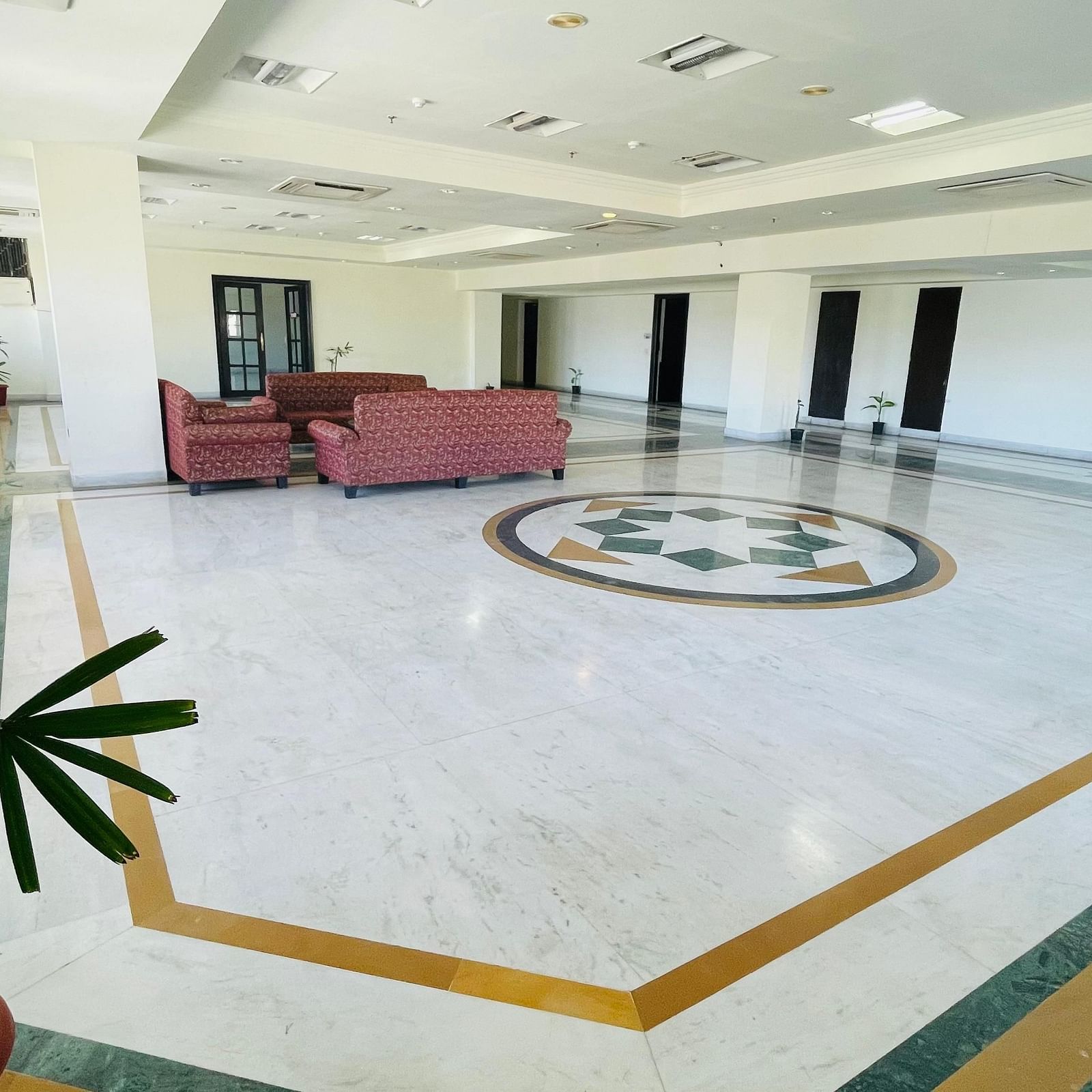 A side view of Raj Vilas Hall featuring potted plants evenly spaced around the tiled floor with sofas at the background.