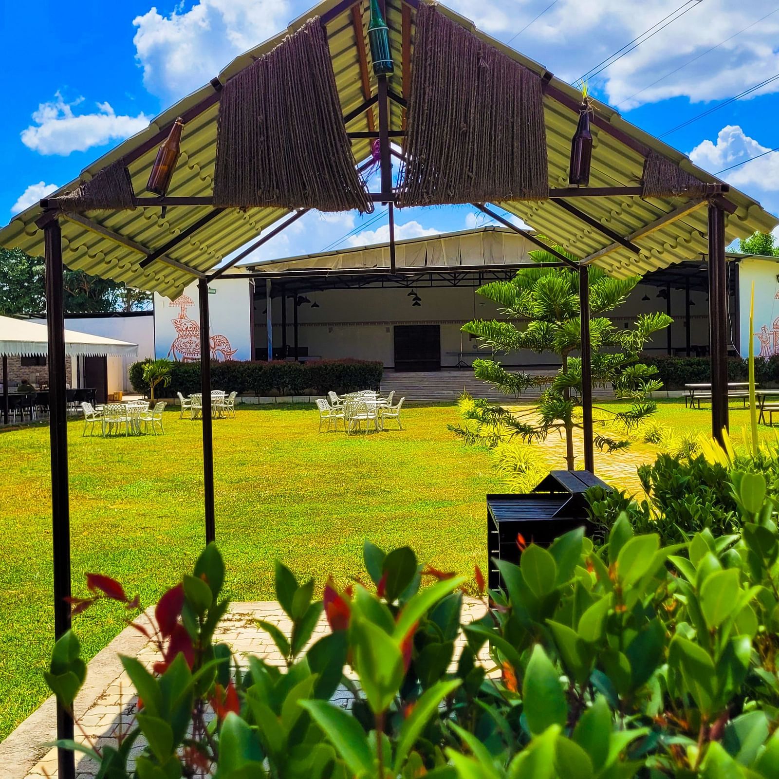 Covered area with tables lined under a beige canopy amidst lush greenery. | Vistar Resort, Bengaluru