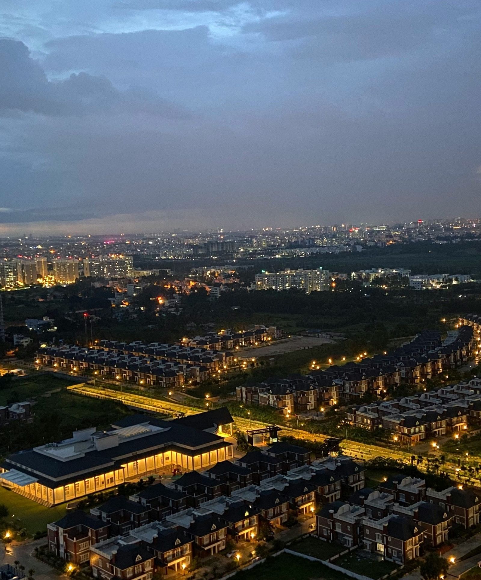 An aerial view of a cityscape at dusk with lights, buildings, and a cloudy sky.