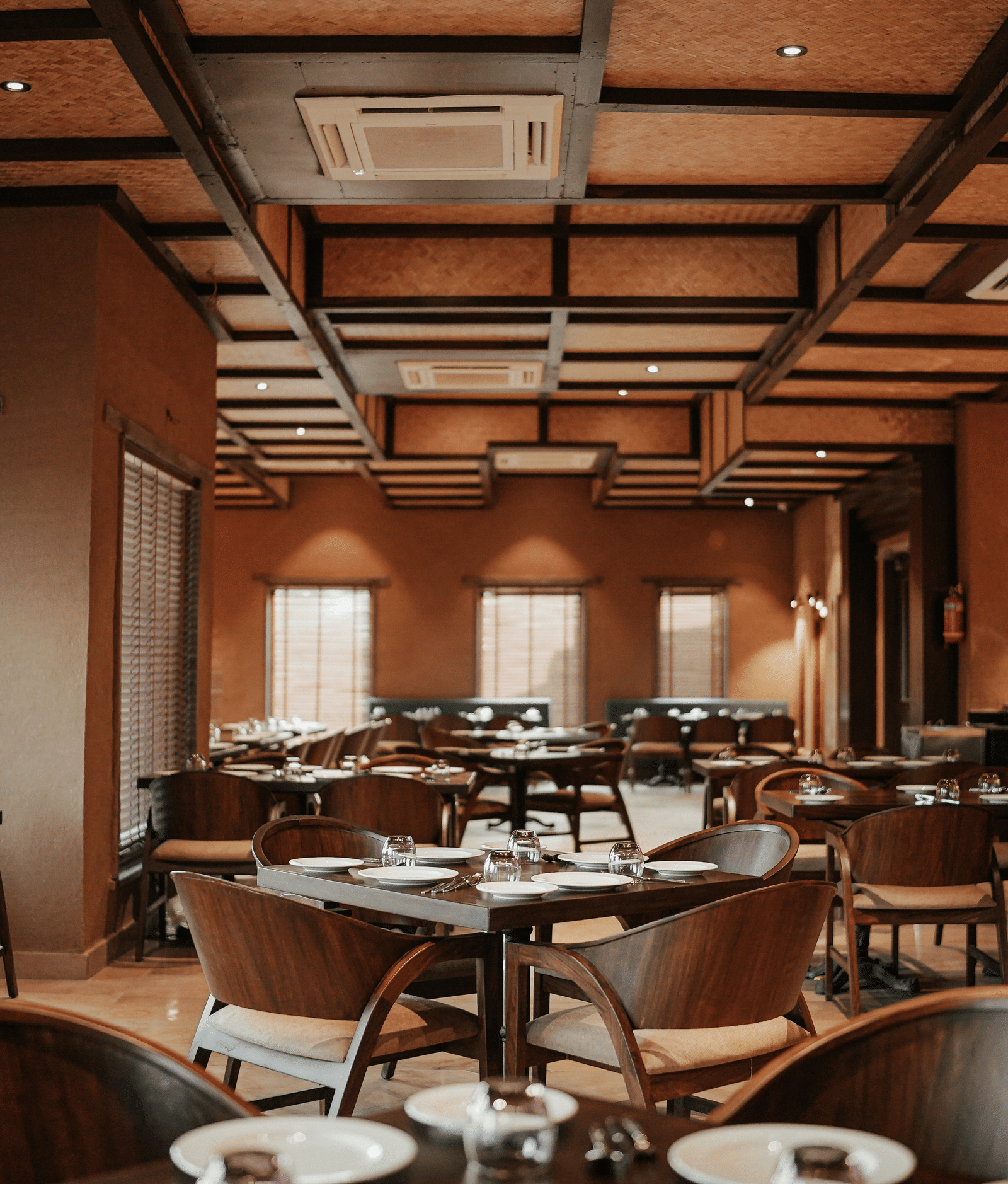 A spacious restaurant hall at Aamaghati Wildlife Resort with wooden ceilings, soft lighting, and neatly arranged tables offA spacious restaurant hall at Aamaghati Wildlife Resort with wooden ceilings, soft lighting, and neatly arranged tables offering gues