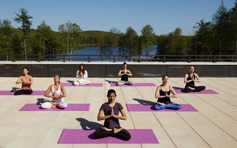 A group of people seated on yoga mats in various meditation poses on an outdoor terrace with a view of a lake and forest.