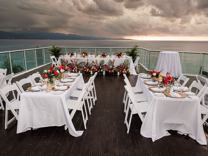 An outdoor dining setup on a terrace with a view of the ocean and a stormy sky. White tables are set with flowers and place settings, and white chairs are arranged around them. The terrace overlooks the sea, and the sky is filled with dark clouds.
