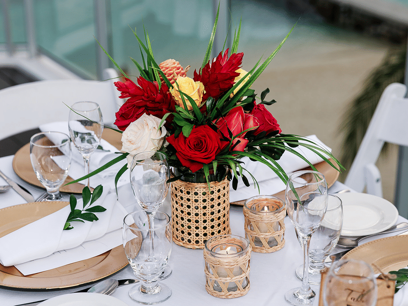 Elegant table setting with a vibrant floral centrepiece of red, yellow, and white roses in a woven vase, surrounded by wine glasses, candles, and neatly arranged place settings.