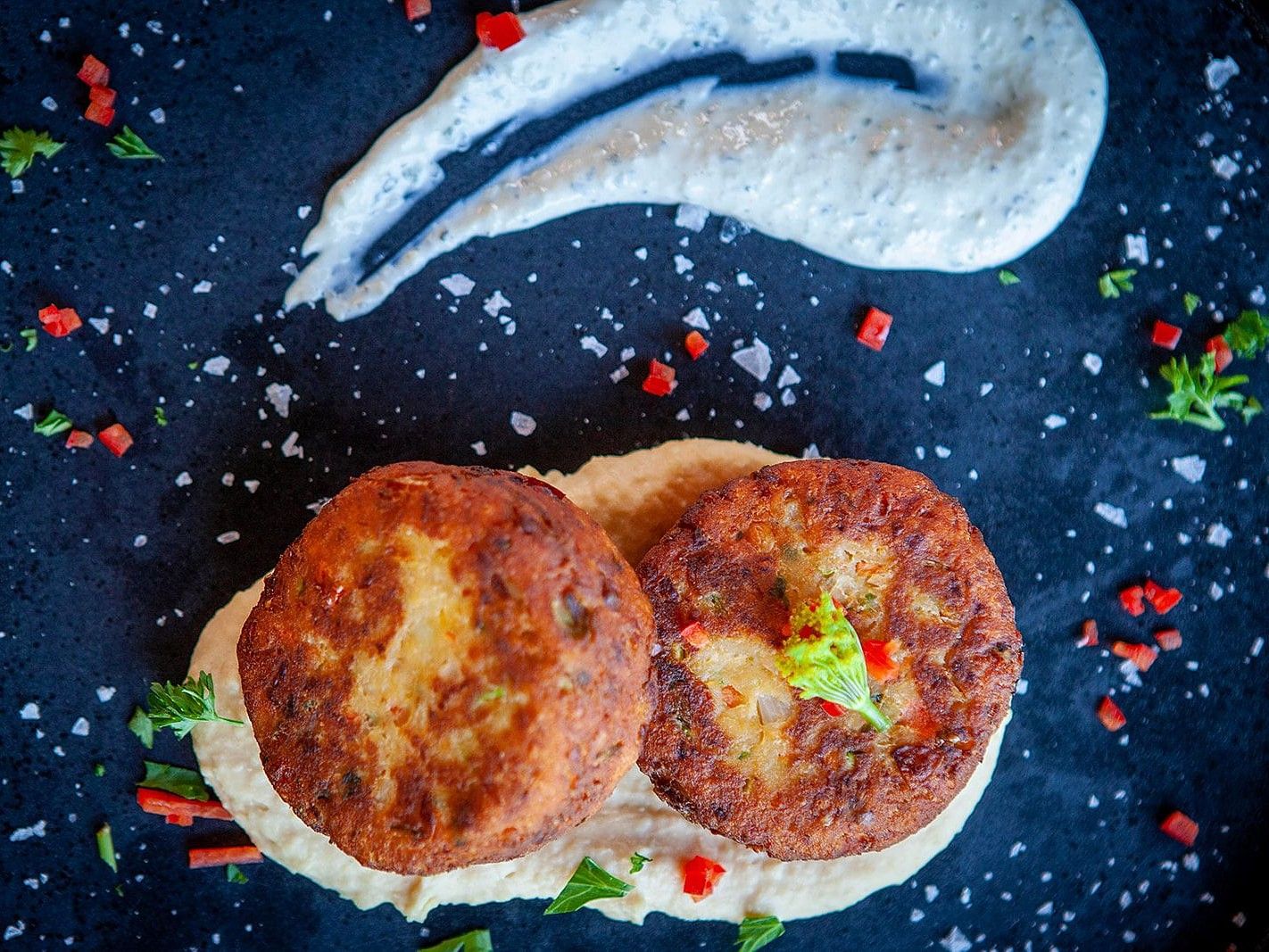 Two golden-brown vegetable patties served on a creamy sauce, garnished with herbs, red peppers, and coarse salt on a black plate at YO1 Longevity and Health Resorts, Catskills.