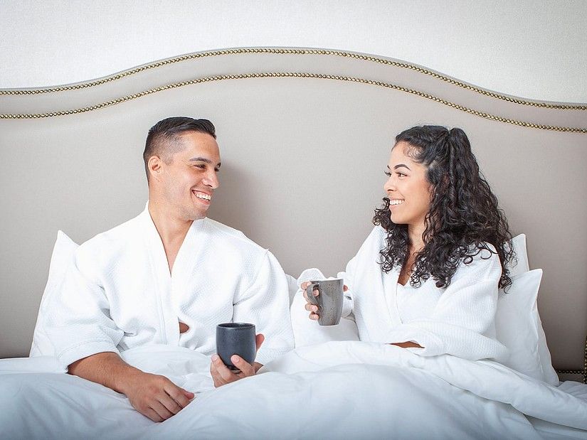 a couple sitting together on a bed under the blanket with coffee mugs in hand