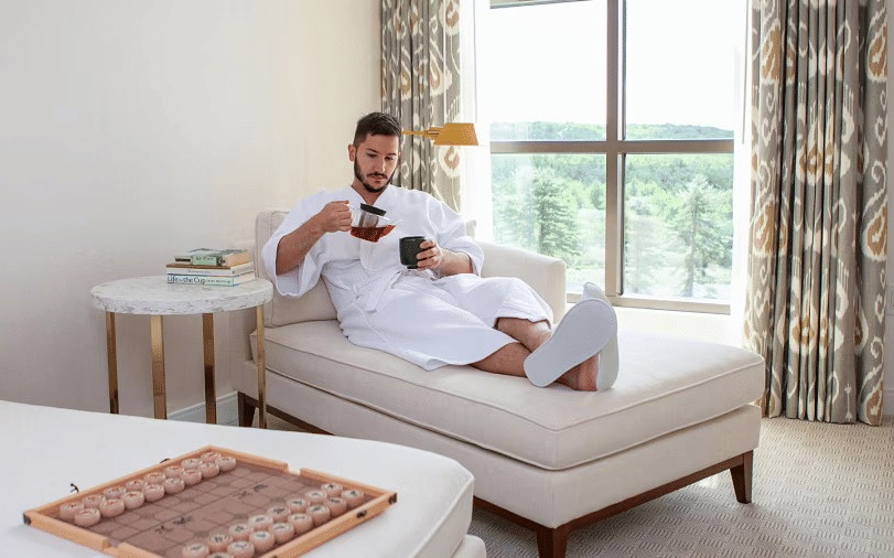  A man in a robe sips from a cup while relaxing on a chaise lounge in a bright room with a large window at YO1 Longevity & Health Resorts