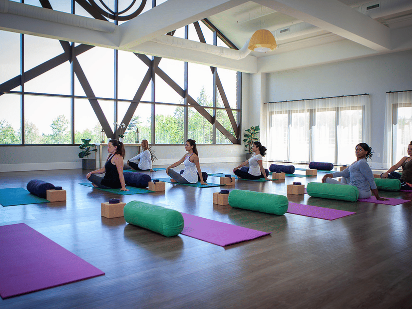 A group of people are practicing yoga in a bright studio with large windows and various props like mats and bolsters at YO1 Longevity and Health Resorts