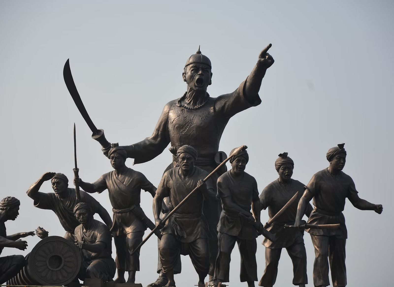 A statue of Lachit Borphukan with soldiers, holding a sword and pointing, against a clear sky.