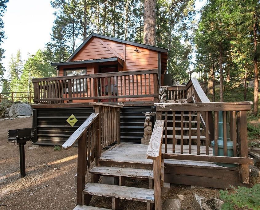 A Cabin at Shaver Lake Village Hotel with a wooden deck, stairs, and a grill, surrounded by tall pine trees.