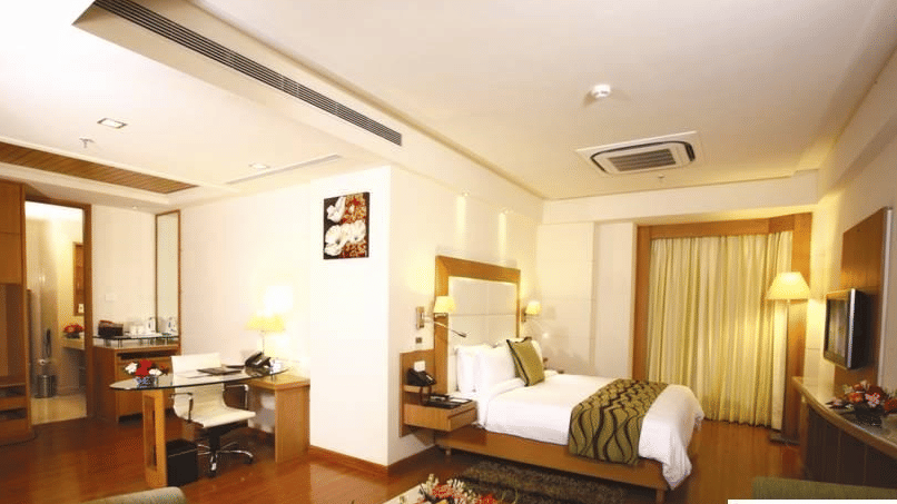  A Premium Suite at Ambrosia Sarovar Portico, Haridwar, with a bed, lounge seating, writing desk, television, and a well-lit open layout.