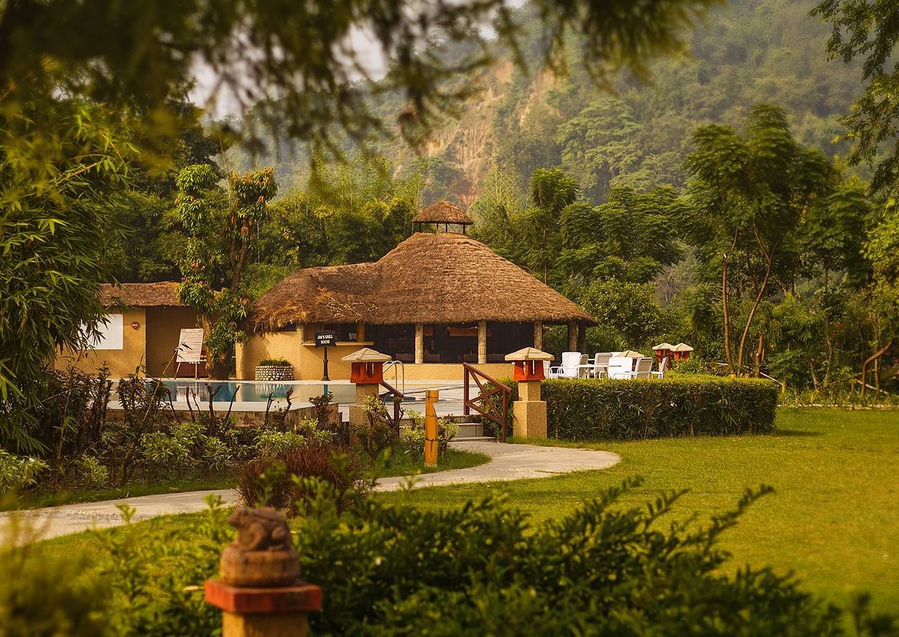 An image of a hut amidst the garden, behind it there is a outdoor seating area with chairs and tea table- The Riverview Retreat, Corbett