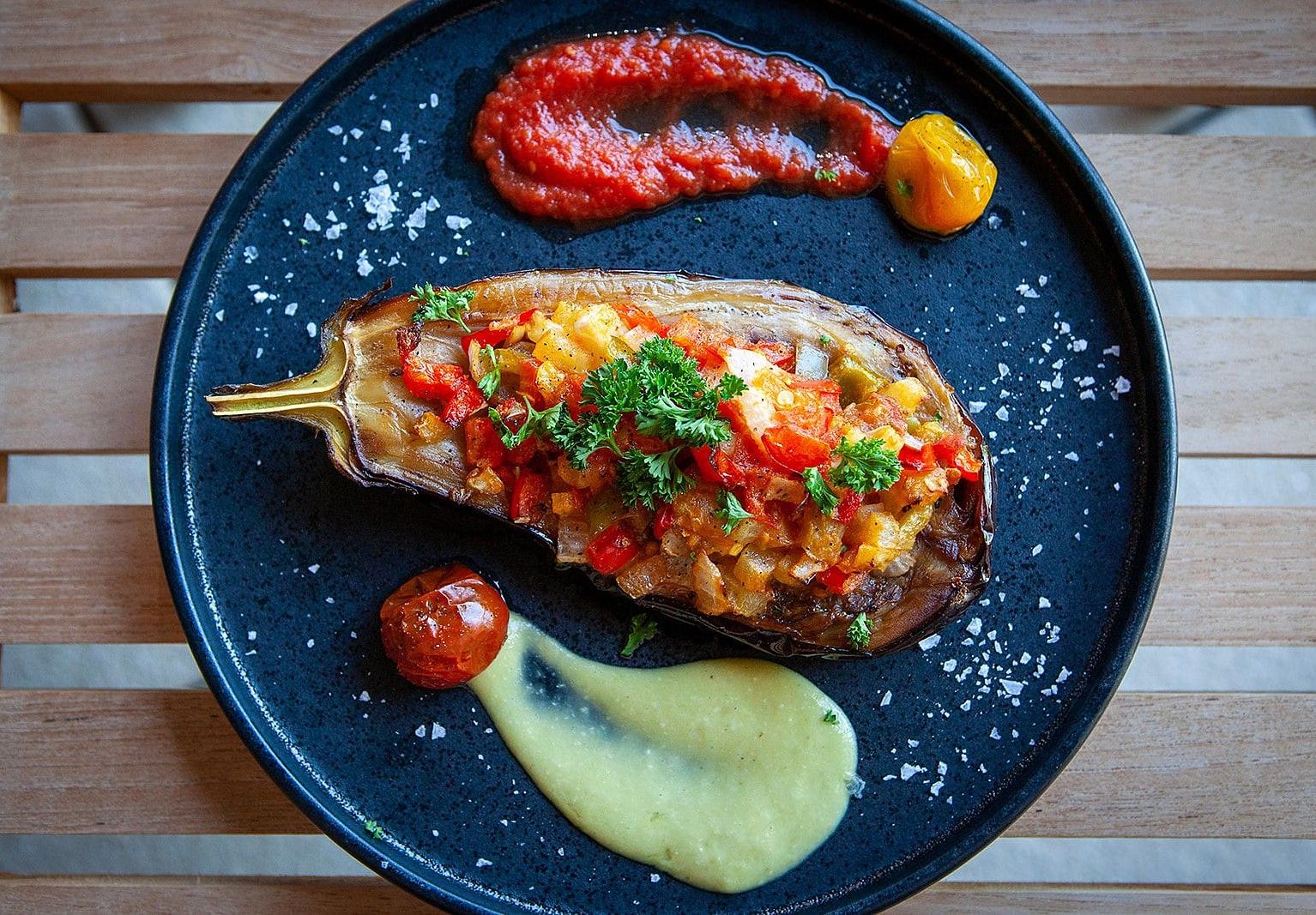 A stuffed eggplant on a black plate accompanied by a swipe of red sauce and a dollop of yellow sauce, garnished with a sprig of parsley at YO1 Longevity & Health Resorts
