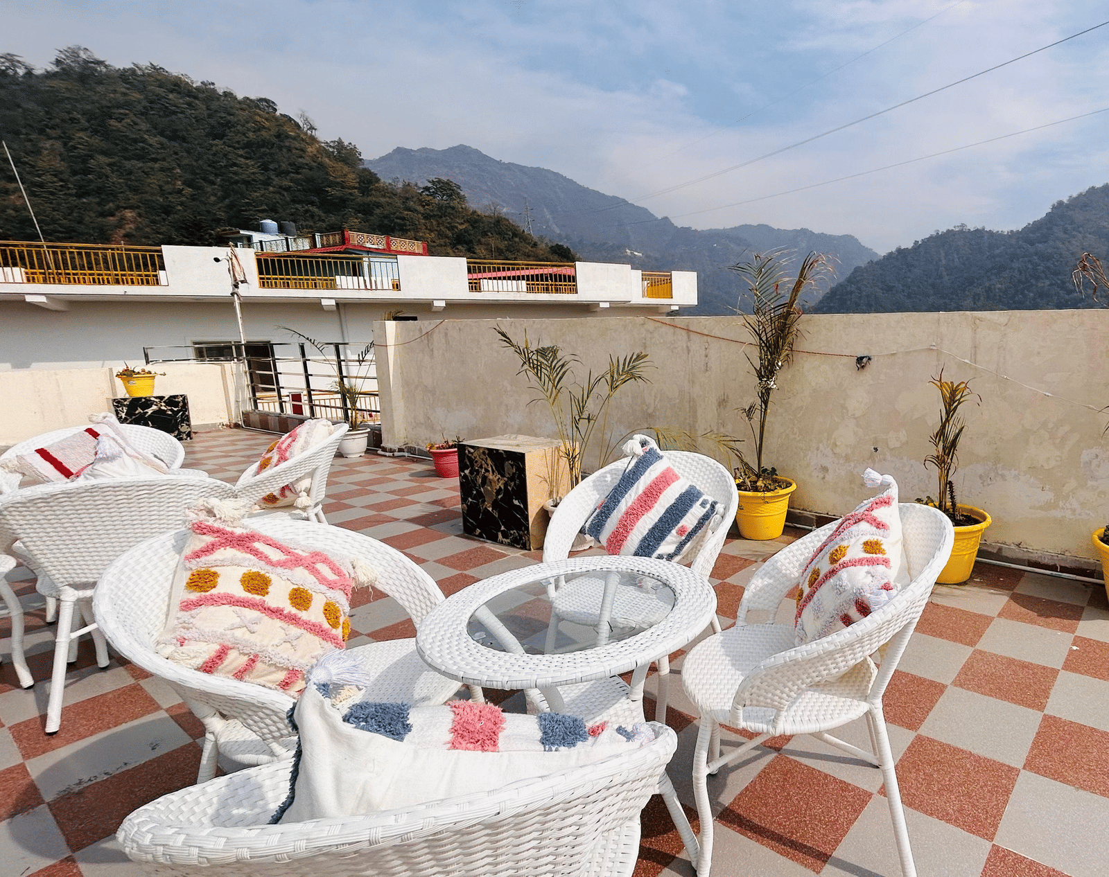 An outdoor rooftop terrace with white wicker tables and chairs and pink accent pillows, overlooking a mountainous landscape | Perfectstayz Value Hills