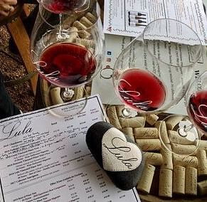 Wine glasses filled with red wine rest on a table with menus, corks, and a heart-shaped object