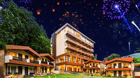 A vibrant night view of DLS Hotels, featuring beautifully lit cottages, a central multi-storey building, lush lawns, and colourful fireworks illuminating the sky.