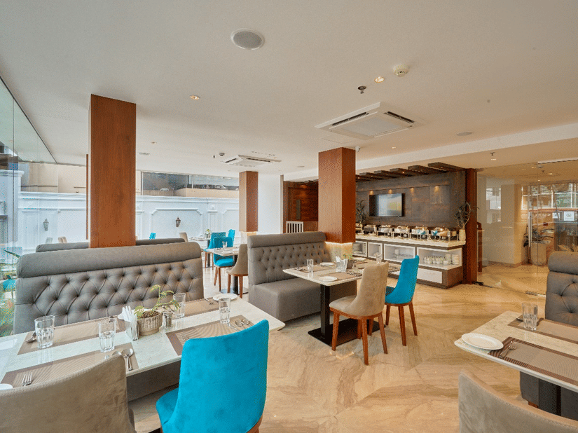 Wide view of the restaurant at QWhite Crescent Luxury Hotel by Dimora Kochi with teal chairs, cushioned booths, and warm ambient lighting.