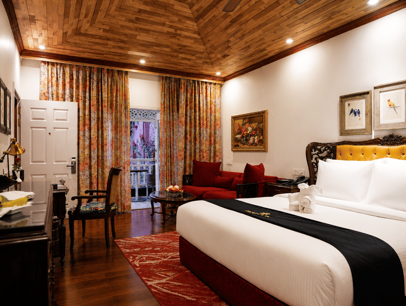 Deluxe Cottage at MAYFAIR Himalayan Spa Resort Kalimpong featuring a bed in the foreground. A sofa sits near a door that opens to the outside. Framed pictures hang on the walls. The ceiling is clad in wood.