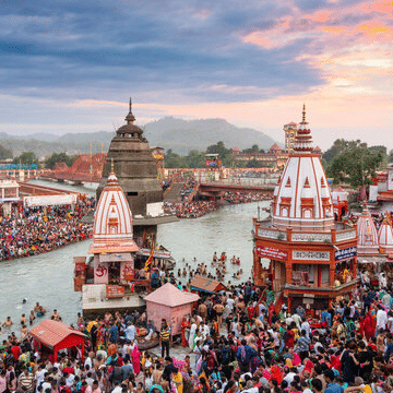 A vast crowd of people gathers near temples along the riverbank under a morning sky.
