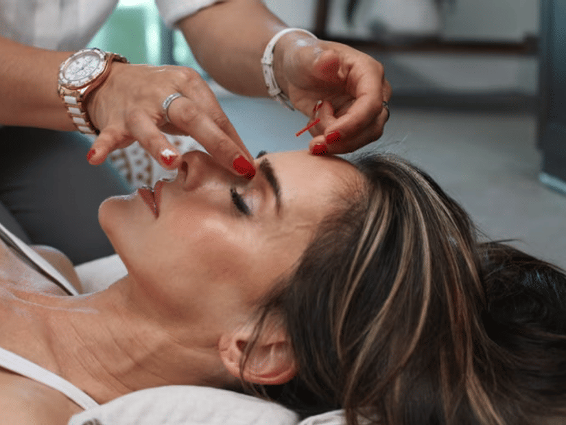 A woman lying back with her eyes closed while a practitioner treats her forehead