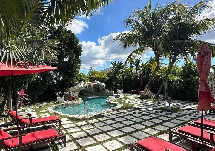 A luxurious backyard pool area with red lounge chairs, umbrellas, and lush palm trees under a bright, partly cloudy sky at A Stone's Throw Away.