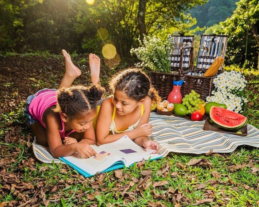 Two girls lying under the sun on a mat with picnic spread on the side and smiling while reading a book