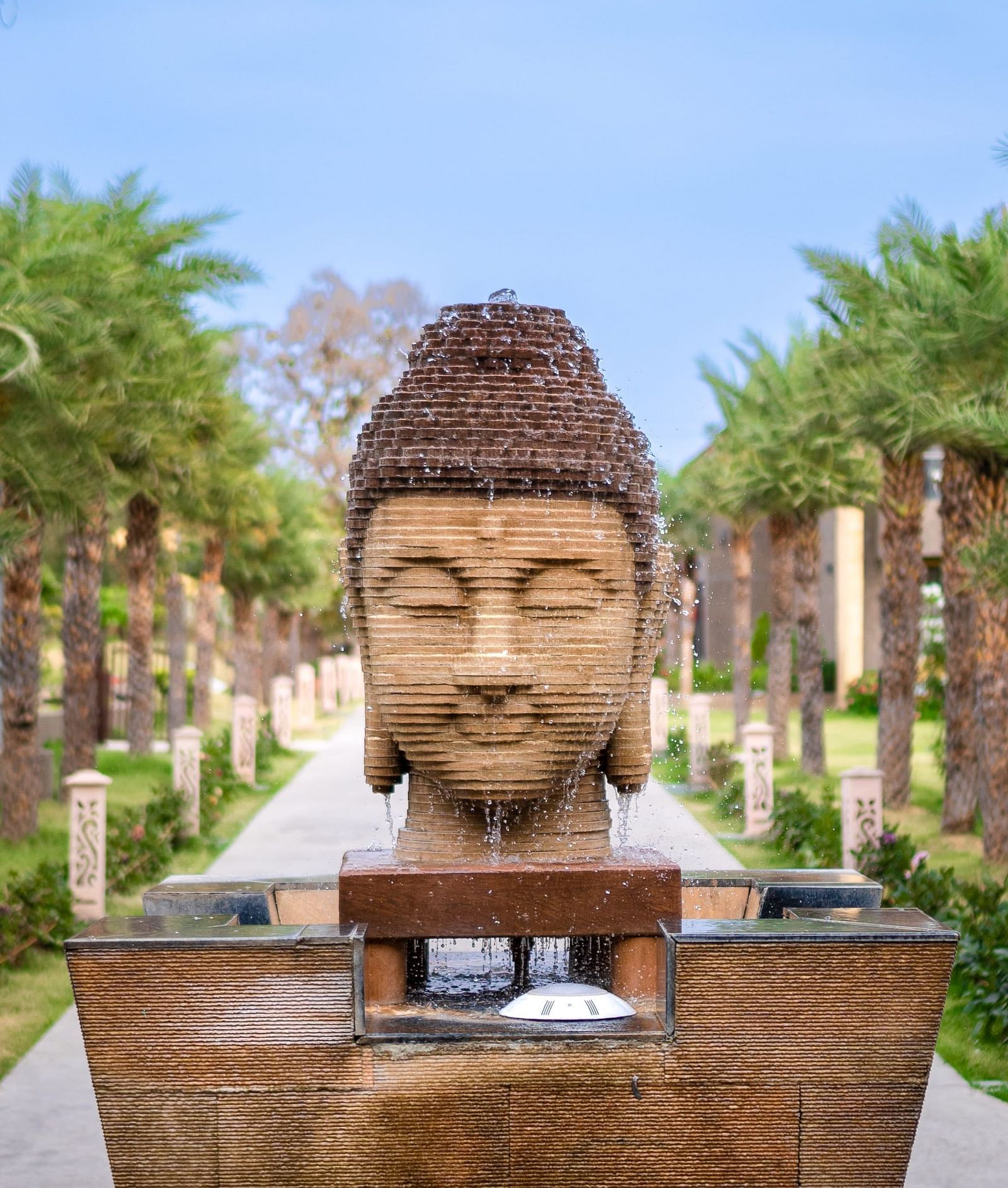 A head bust of Buddha on a pedestal, in the middle of a walk path, which has pal trees on either side - Crescent Resorts