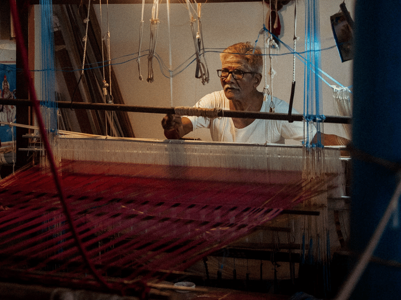 An elderly artisan operates a traditional handloom inside a weaving workshop, crafting vibrant fabric using time-honoured techniques and skilled craftsmanship.