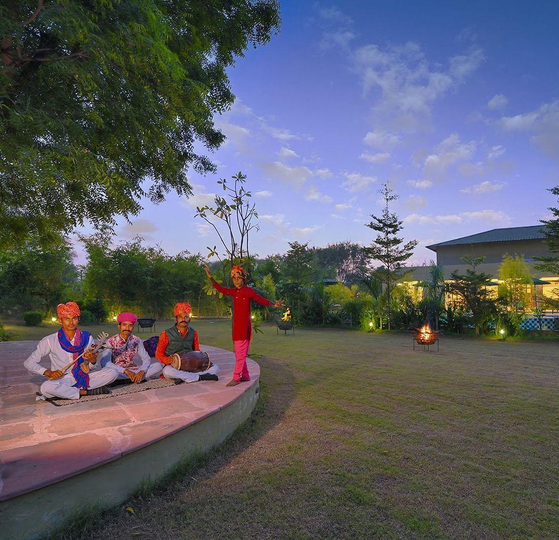 Three musicians in traditional attire playing instruments on a stone platform in front of a resort building at dusk - Singhvilas, Ranthambore