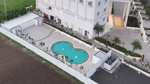 A multi-story white building of Pride Resort, Rajkot featuring a swimming pool, surrounded by green lawns and undeveloped land