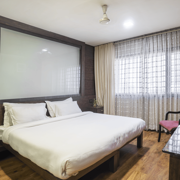 A spacious room with a king size bed, beige curtains, dark wall panelling, a pink armchair, and a T.V. at Infiniti Hotel, Indore.