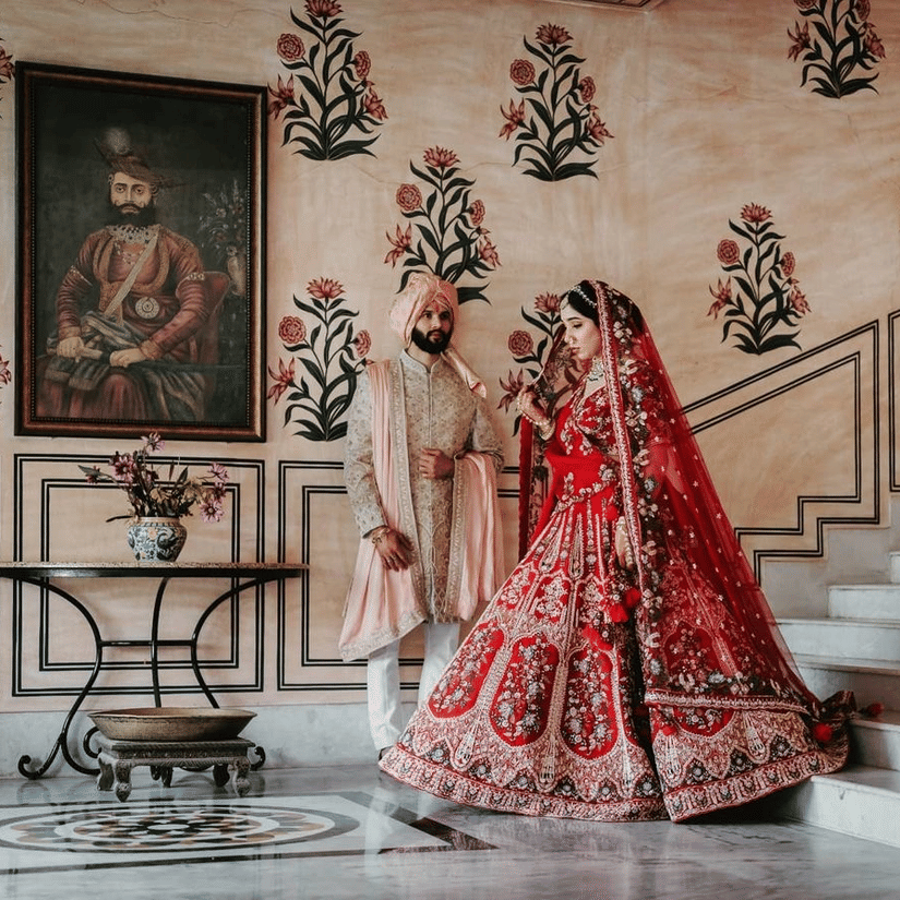 A bride and groom stand together indoors, with a portrait on the wall and floral designs behind them at  Umaid Palace - Best Luxury Heritage Resort Near Jaipur.  
