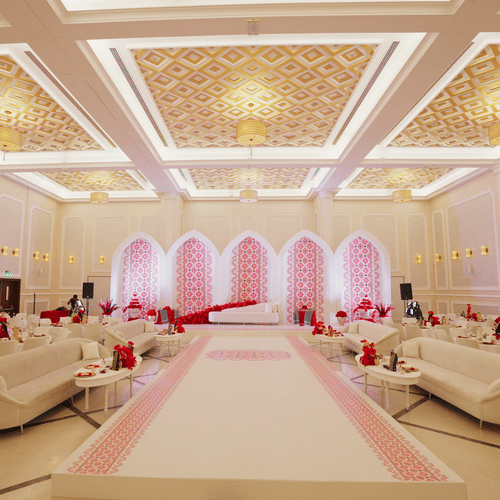 A luxurious wedding hall with elegant chandeliers, white and gold decor, and soft pink lighting highlighting an aisle lined with plush sofas. The setting is opulent and designed for an extravagant event - La Maison Hotel, Doha