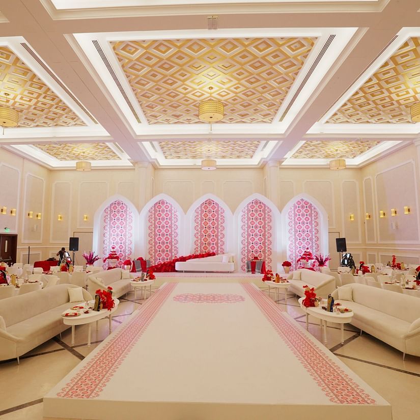 Luxurious wedding hall with elegant chandeliers, white and gold decor, and soft pink lighting highlighting an aisle lined with plush sofas. The setting is opulent and designed for an extravagant event - La Maison, Doha
