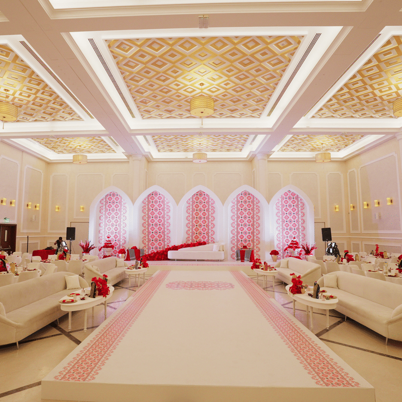 A luxurious wedding hall with elegant chandeliers, white and gold decor, and soft pink lighting highlighting an aisle lined with plush sofas. The setting is opulent and designed for an extravagant event - La Maison Hotel, Doha