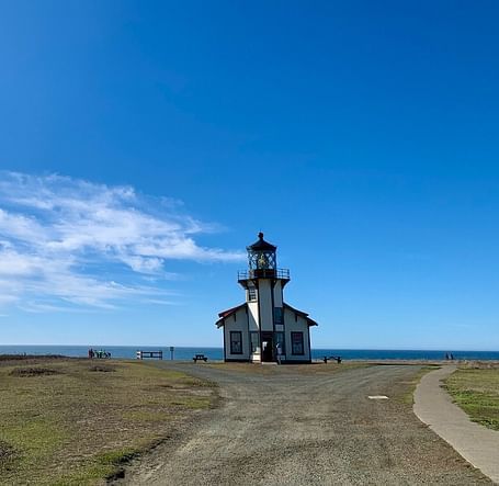 A gravel path leads to a lighthouse perched on a grassy hill overlooking the ocean under a vast sky.