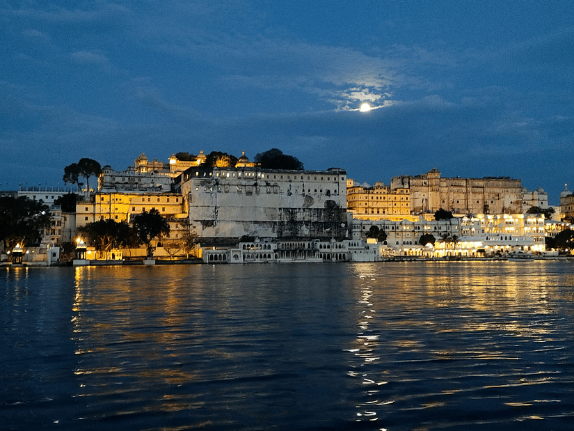 A view from afar of the City Palace with Lake Pichola in front of it and the moon shining above in the sky at Udaipur Ambrai Ghat.